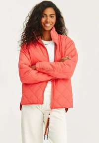 Koralowa pikowana kurtka z wysokim kołnierzem, zamkiem z przodu i kieszeniami bocznymi. Dekoracyjne akcenty w postaci sznureczków na dole. Widoczna biała warstwa wewnętrzna.
