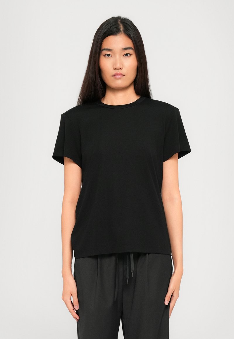 Young woman with long dark hair wearing a plain black short-sleeve t-shirt and dark pants, standing against a white background.