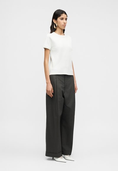 Λευκό τοπ με κοντά μανίκια συνδυασμένο με σκούρο, ψηλόμεσο παντελόνι φαρδιάς γραμμής. Το μοντέλο φορά έντονα χρυσά σκουλαρίκια κρίκους και λευκά μυτερά παπούτσια.