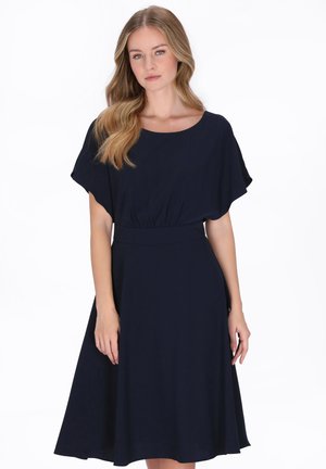 Fiatal nő hosszú, hullámos szőke hajjal, térdig érő sötétkék ruhában, rövid ujjú, fehér háttér előtt áll.