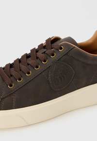 Brown leather sneaker with round toe, gold eyelets, and dark brown laces. Features a beige rubber sole and a shield logo on the side.