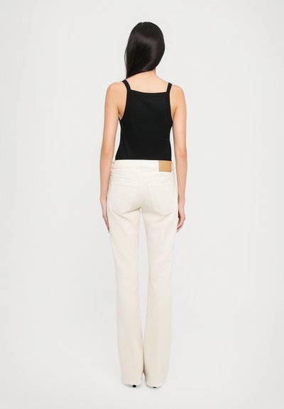 Mujer con cabello largo y oscuro, vistiendo un top sin mangas negro y unos jeans rectos de talle alto en color blanco roto, de pie contra un fondo blanco.