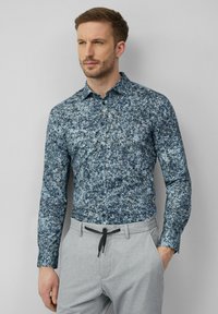 Chemisier bleu foncé avec un imprime floral en bleu clair et tons crème ; il possède des manches longues et un col pointu, associé à un pantalon gris clair.