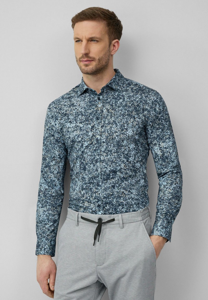 Chemisier bleu foncé avec un imprime floral en bleu clair et tons crème ; il possède des manches longues et un col pointu, associé à un pantalon gris clair.