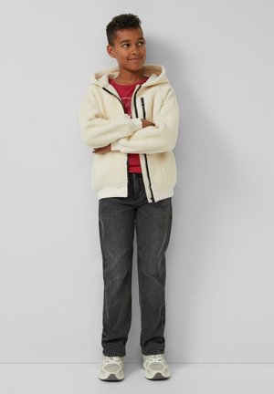 Cremfarbener Fleece-Hoodie mit Reißverschluss, schwarzen Akzenten, rotem T-Shirt darunter, dunkler Jeans und hellen Sneakern. Arme gekreuzt.