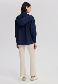 Tamno plava dukserica s kapuljačom, s napućenim rukavima i ležernim krojem, nosi se preko krem boje širokih hlača i bijelih tenisica, prikazuje glatku teksturu.