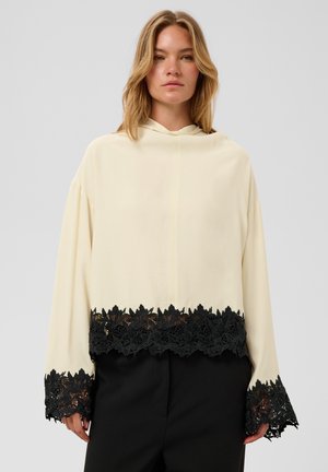 Vrouw draagt een crèmekleurige top met lange mouwen en zwarte bloemkant op de mouwen en zoom, gecombineerd met een zwarte broek, staand tegen een effen achtergrond.