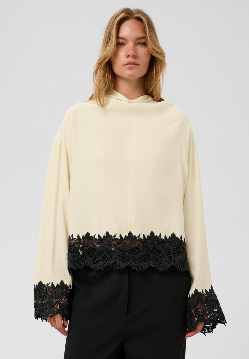 Femme portant un haut crème à manches longues avec une bordure en dentelle florale noire aux manches et à l'ourlet, associé à un pantalon noir, debout devant un fond uni.