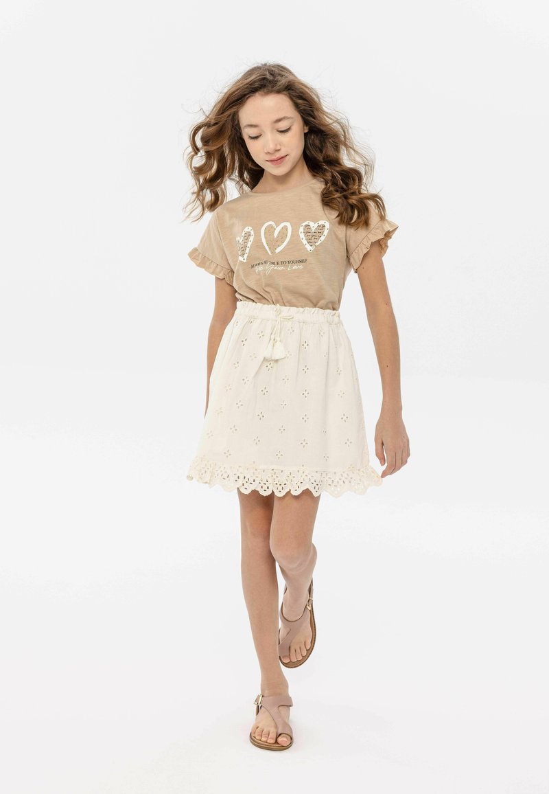 Chemise marron à manches courtes avec un motif de cœur blanc, associée à une jupe blanche en dentelle avec un ourlet festonné. Des sandales beiges complètent la tenue.
