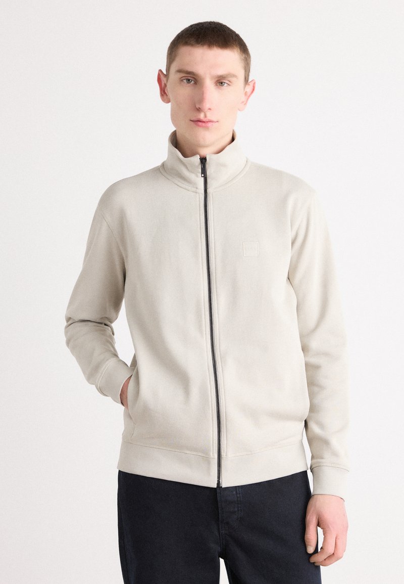 Sweatshirt bege claro com fecho éclair, gola alta, punhos canelados e um discreto logótipo no peito. Tecido suave, corte descontraído.
