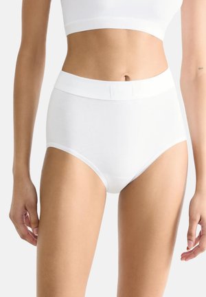 Femme portant un slip taille haute blanc et un haut court blanc, debout devant un arrière-plan clair uni.