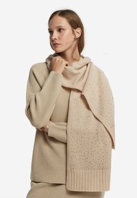 Beżowy dziergany szal z teksturowanym wzorem i subtelnymi ozdobami, zarzucony na ramiona, dopełniający pasujący sweter.