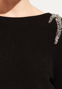 Pull en tricot côtelé noir avec un décolleté large et des embellissements décoratifs en argent sur l'épaule. Le détail du tissu texturé est visible.