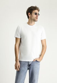 T-shirt bianca a maniche corte in cotone liscio, abbinata a jeans in denim azzurro chiaro. Il modello indossa occhiali da sole rotondi con montatura sottile.