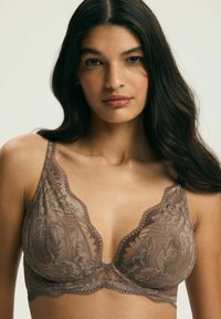 Žena s dlhými tmavými vlasmi v hnedom čipkovom podprsenke, čelom k fotoaparátu s neutrálne výrazom, jednoliate svetlé pozadie.