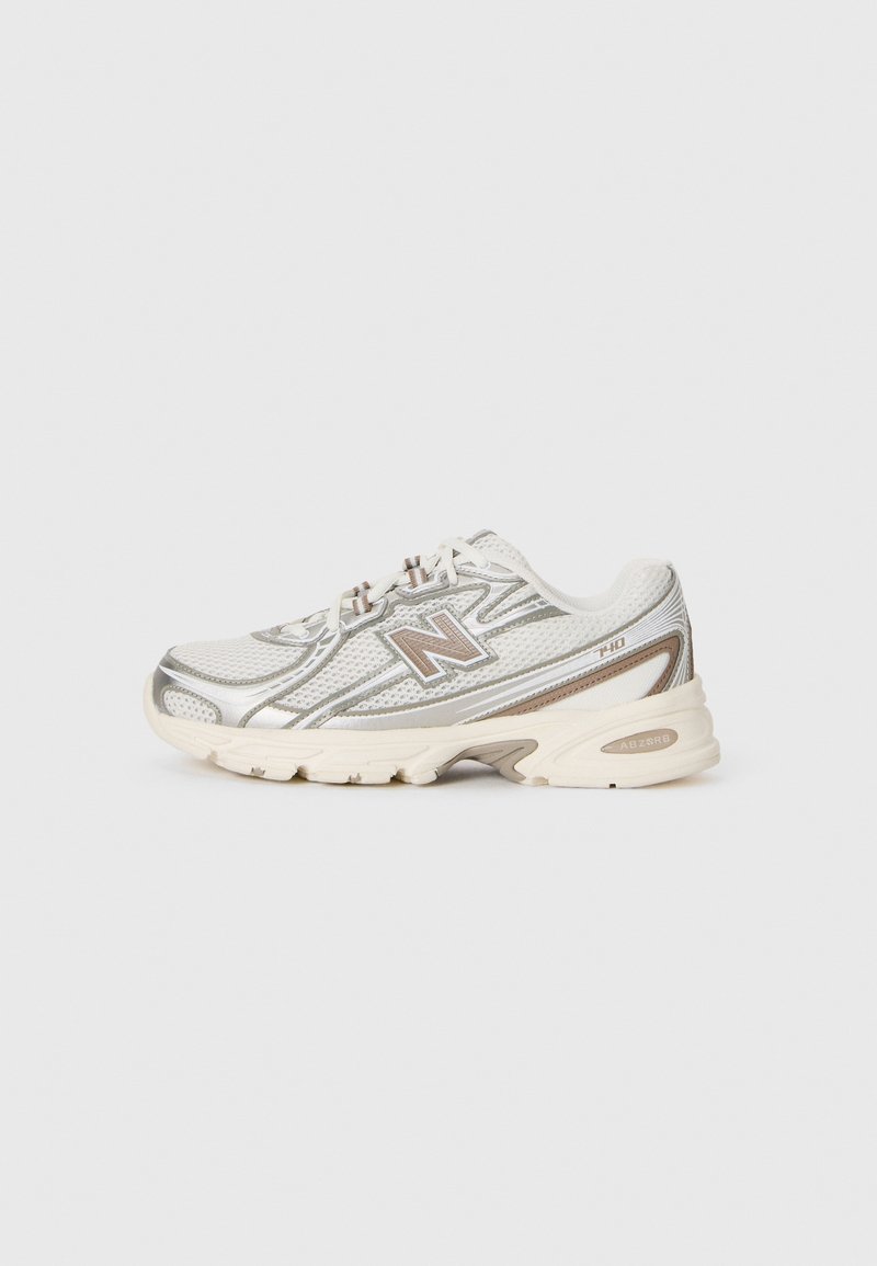 Chaussure de course New Balance blanche et argentée avec des accents beiges, tige en mesh et semelle rembourrée sur un fond gris clair uni.