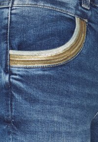 Denimjeans med en bakficka som har ett böjt, guldmetalliskt mönster och ljusblå sömmar. Tyget har en bleknad blå textur.