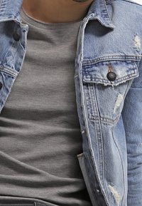 Jeansjacka i ljusblå färg, med slitna detaljer och svarta knappar. Under jackan en grå, strukturerad långärmad tröja.