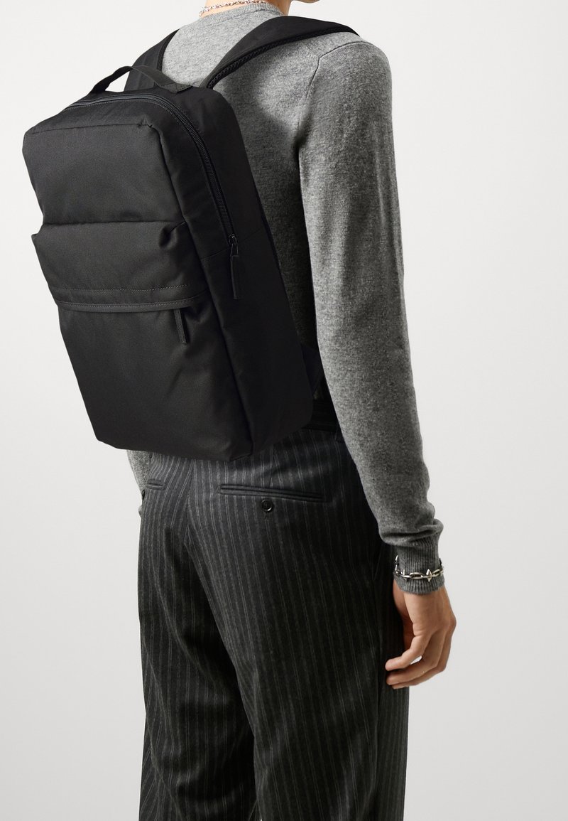 Personne portant un sac à dos rectangulaire noir avec des poches zippées, un t-shirt gris à manches longues et un pantalon sombre à fines rayures sur fond blanc.
