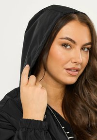 Giacca nera con cappuccio, in tessuto liscio, con polsini elastici e zip frontale, che presenta un modello che regola il cappuccio.