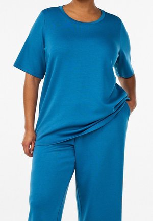 Personne portant un haut bleu ample à manches courtes et un pantalon large assorti, une main dans la poche du pantalon.