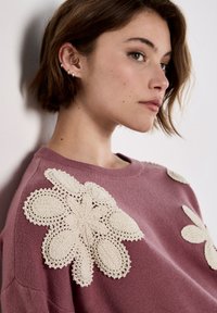 Rosa Sweatshirt mit großen, filigranen beigen Häkelblumen-Applikationen auf den Schultern, runder Ausschnitt und strukturiertem Stoff.
