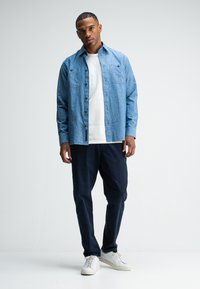 Lichtblauwe overhemdd met twee borstzakken, gecombineerd met marineblauwe broeken en witte sneakers. Soepele stof; casual pasvorm.