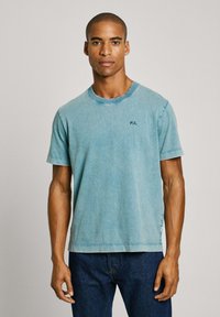 Světle modré denimové tričko s kulatým výstřihem, krátkými rukávy a bočními rozparky. Na hrudi je vyšitý nápis "PJL" tmavě modrou barvou.