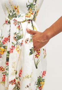 Vestido de tela blanca con un estampado floral, que presenta una cintura atada y un bolsillo lateral, con flores de colores en tonos de rosa, amarillo y verde.