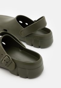 Zoccoli di gomma verde scuro con forma arrotondata, cinturino posteriore regolabile e fori di ventilazione. Presentano il logo del marchio in rilievo e una suola testurizzata.