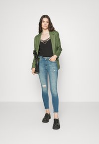Blazer a cuadros verdes, camisón de encaje negro y jeans ajustados desgastados de color azul claro. Mocasines de plataforma negros. Fondo simple.