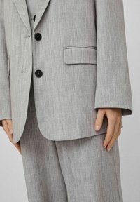 Ljusgrå skräddarsydd kostym med en texturerad yta. Har svarta knappar, en enda bröstficka och vida ärmar.