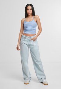 Vrouw met lang, donker haar, gekleed in een blauw-wit gestreept tanktop, lichtblauwe wijde jeans en beige sneakers, tegen een witte achtergrond.