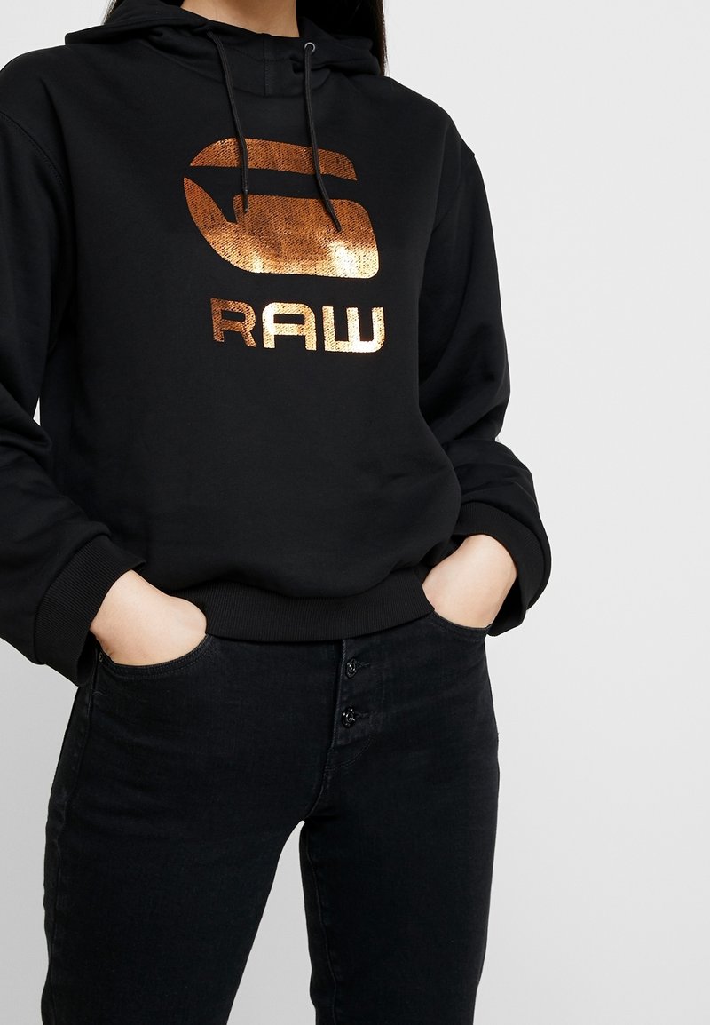 Persona vestida con una sudadera con capucha negra y un logo "RAW" de color cobre, con las manos en los bolsillos, combinada con unos vaqueros de tiro alto negros con botones.