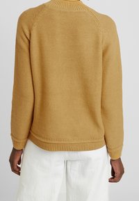 Pull moutarde avec un tricot texturé, un col rond et un ourlet côtelé. Comprend des manches raglan et une coupe décontractée. Pantalon blanc visible en dessous.