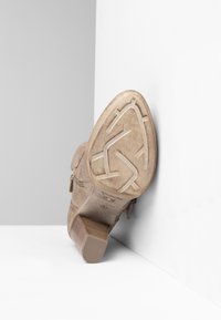 Botín de ante beige con suela texturizada, leve tacón y detalle de cremallera lateral, posicionado sobre un fondo blanco.