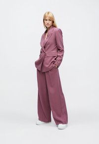 Rosa maßgeschneiderter Anzug mit einem strukturierten, doppelt geknöpften Blazer und weit geschnittenen Hosen, kombiniert mit weißen Sneakern. Glatte Textur, minimalistische Details.