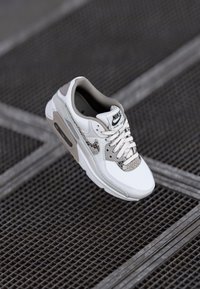 Nike Air Max 90 apavi ar baltu ādas augšdaļu, pelēkiem akcentiem, sietu uzliktu un strukturētu čūskas rakstu detaļu uz pirksta.