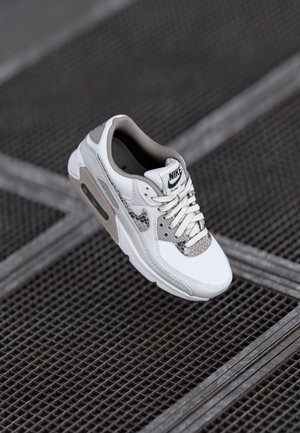 Adidașii Nike Air Max 90 au un superior din piele albă cu accente gri, limbă din plasă și detalii texturate cu model de șarpe pe vârf.