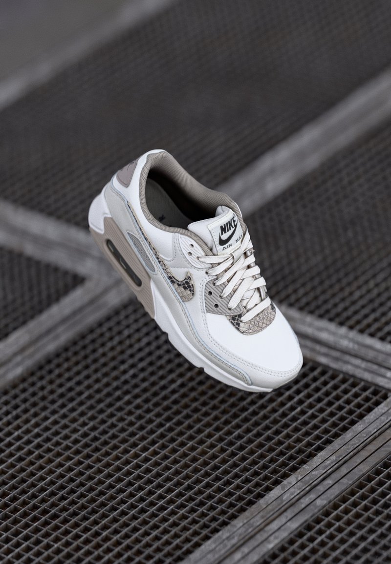 Nike Air Max 90 apavi ar baltu ādas augšdaļu, pelēkiem akcentiem, sietu uzliktu un strukturētu čūskas rakstu detaļu uz pirksta.