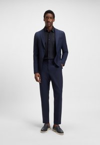 Marineblaues Anzug mit Revers, einreihiger Jacke und schlanken Hosen. Über einem schwarzen Hemd getragen, kombiniert mit dunklen Slippers.