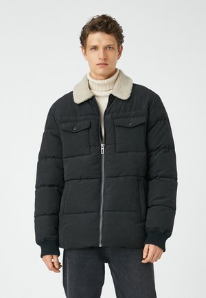 Veste noire matelassée avec col en peau lainée beige, fermeture éclair à l'avant et deux poches poitrine. Poignets côtelés et silhouette droite.