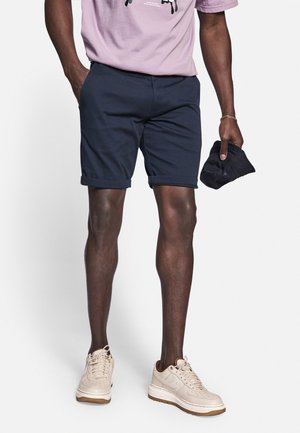 En person iført marineblå op-rullede shorts, lyse beige sneakers og en lilla T-shirt, der holder en marineblå kasket i højre hånd.