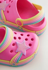 Helle pinke Clogs mit regenbogenfarbenen Riemen und sternförmigen Verzierungen, die Belüftungslöcher auf der Oberseite aufweisen.