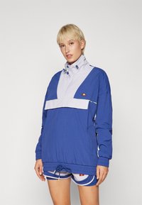 Junge Person mit kurzem blonden Haar, die eine blaue und helllila Windjacke sowie passende Shorts trägt, vor einem einfarbigen Hintergrund.