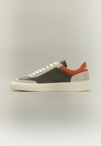 Baskets basses en cuir vert foncé, accents en daim beige, lacets blancs, détail rouge au talon et semelle couleur crème sur un fond uni.