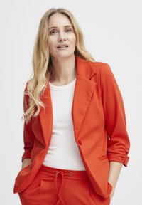 Červený přizpůsobený blazer se strukturovaným designem, zapínání na jeden knoflík a ohrnutými rukávy. Spárován s bílým upnutým topem, což vytváří soudržný outfit.