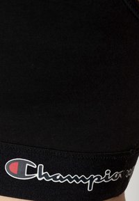 Czarny stanik sportowy wykonany z elastycznego materiału, z szerokim elastycznym pasem z logo "Champion" w białych i czerwonych akcentach.