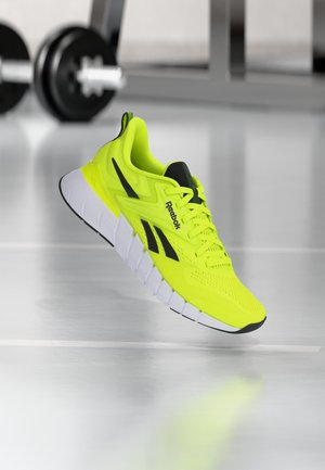 Ryškiai geltona Reebok sportinė avalynė su juodais akcentais ir balta pado dalimi, sklandanti virš blizgančios sporto salės grindų, fone matosi svoriai.