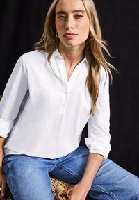 Chemise blanche à manches longues avec un col décontracté et une patte de boutonnage, associée à un jean en denim bleu. Texture lisse, design simple, coupe décontractée.